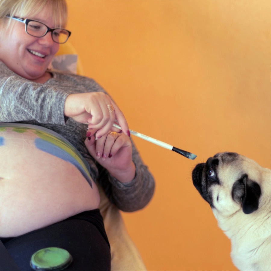 Lachende Schwangere spielt mit ihrem Mops, der neugierig am Pinsel der Babybauchbemalung schnuppert