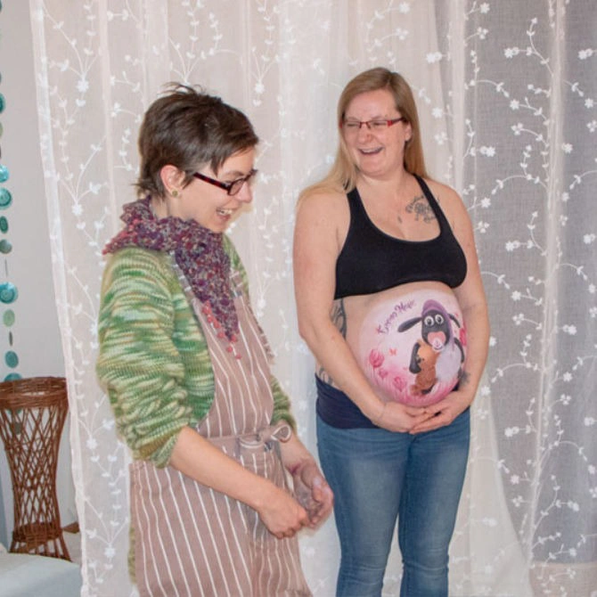 Künstlerin übt mit der schwangeren Frau eine Pose für das Fotoshooting der Babybauchbemalung mit Shaun das Schaf
