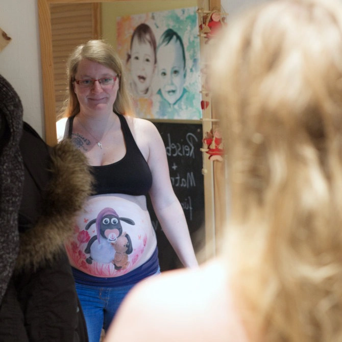 Schwangere Frau betrachtet im Spiegel ihre fertige Babybauchbemalung mit Shaun das Schaf und Teddy