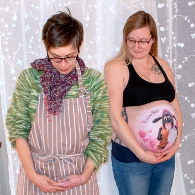 Künstlerin und schwangere Frau stehen nebeneinander und halten stolz die fertige Babybauchbemalung mit Shaun das Schaf und Teddy