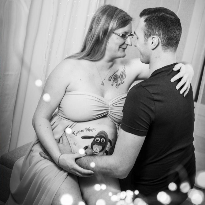 Schwangere Frau mit bemaltem Babybauch im Shaun-das-Schaf-Motiv sitzt liebevoll mit ihrem Partner beim Fotoshooting