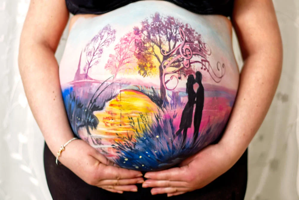 Kundin mit romantischer Babybauchbemalung: Paar bei Sonnenuntergang unter Musikbaum – Testimonial cy-DESIGN