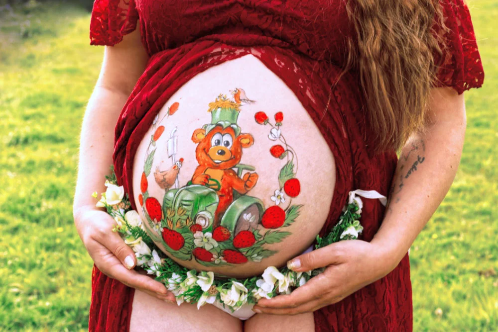 Babybauchbemalung mit Bär auf Traktor, Erdbeeren und Blumen – verspieltes Bauernhof-Motiv auf rotem Kleid im Sommer