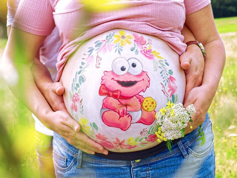 Bunte Babybauchbemalung mit Elmo-Motiv, Blumenranken und liebevoller Umarmung – verspieltes Motiv für werdende Eltern