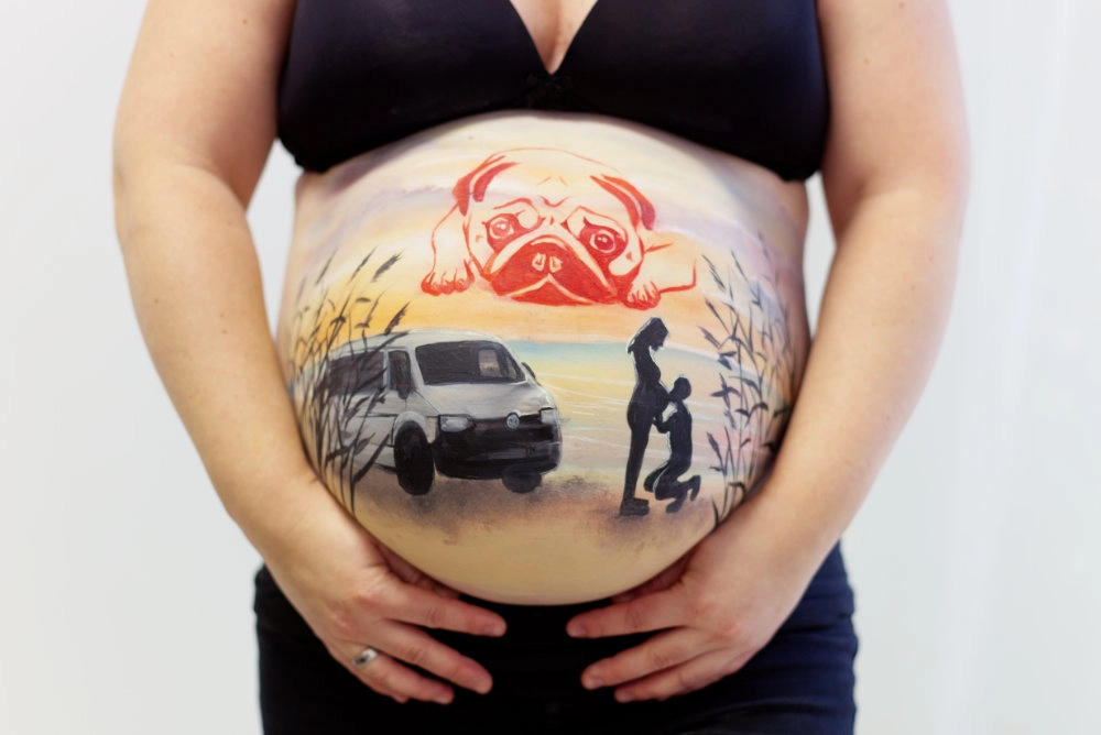 Individuelle Babybauchbemalung mit Mops, Familienauto und Silhouette eines Paares am Strand bei Sonnenuntergang – persönliche Erinnerung mit Humor und Herz