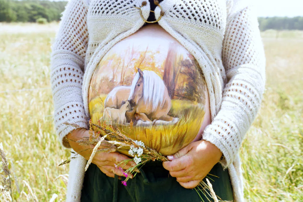 Herbstliche Babybauchbemalung mit ruhender Stute und Fohlen auf einer Waldwiese – naturverbundenes Pferdemotiv als liebevolle Erinnerung an die Schwangerschaft