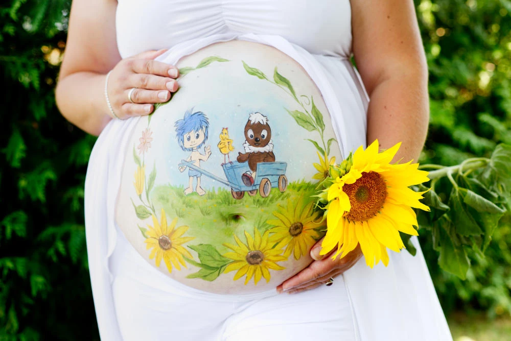 Sommerliche Babybauchbemalung mit Sonnenblumen, zwei fröhlichen Figuren und einer Schubkarre – kindliches, verspieltes Motiv auf grüner Wiese als liebevolle Erinnerung an die Schwangerschaft