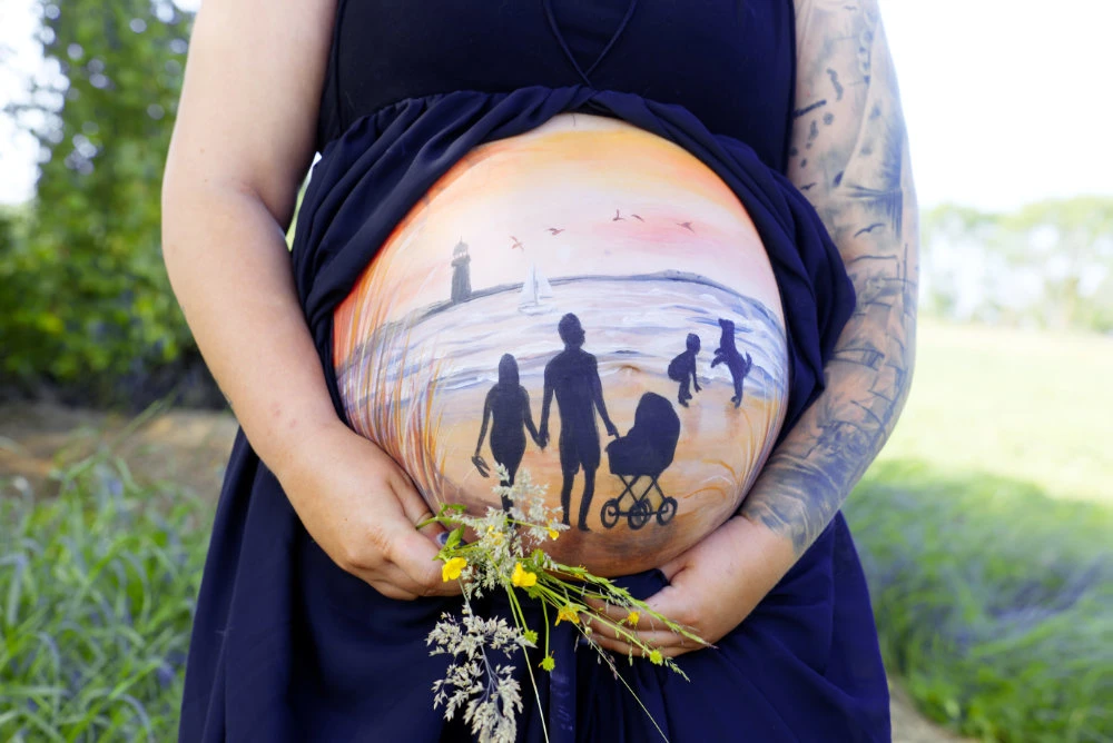 Babybauchbemalung mit Silhouetten einer Familie am Strand bei Sonnenuntergang – emotionales Motiv mit Kinderwagen, Hund und Leuchtturm als Symbol für Zusammenhalt und Vorfreude
