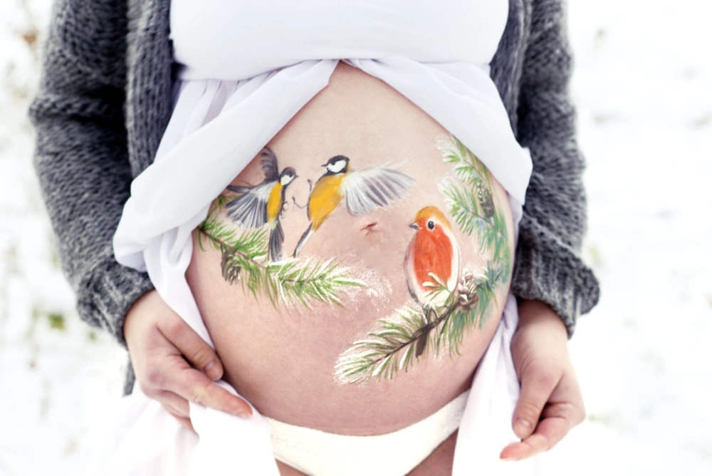 Babybauchbemalung mit Wintermotiv: zwei Meisen und ein Rotkehlchen auf Tannenzweigen – zarte, naturinspirierte Schwangerschaftsbemalung mit Vogel-Illustration
