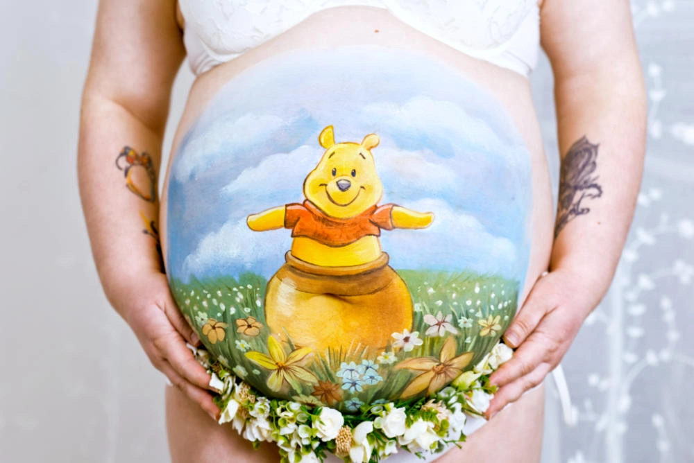 Bunte Babybauchbemalung mit Winnie Puuh auf einer Blumenwiese – verspieltes Motiv für werdende Eltern mit fröhlichem Kinderbuch-Charakter