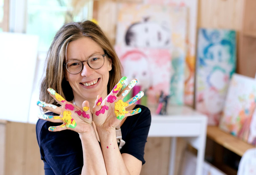 Conny Halliger in ihrem Atelier – lächelnde Künstlerin mit farbenfrohen bemalten Händen vor ihren Bildern, spezialisiert auf Babybauchbemalung und kreative Malerei